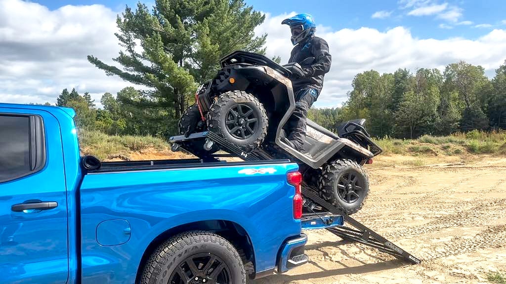 Un homme portant un casque et des vêtements de protection conduit un VTT Segway sur une rampe pour le charger dans le plateau d’un pick-up bleu sur un terrain de sable.