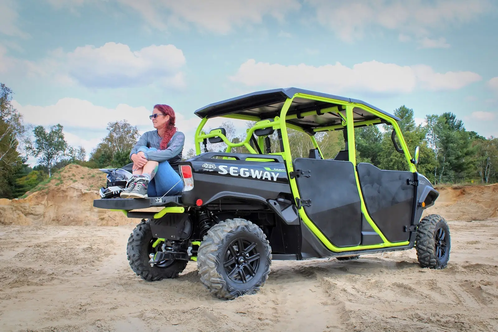 A woman sitting in the back of a Fugleman UT10 Crew, Segway’s six-seater utility side-by-side. The vehicle is parked on a sandy terrain with hills and trees in the background.